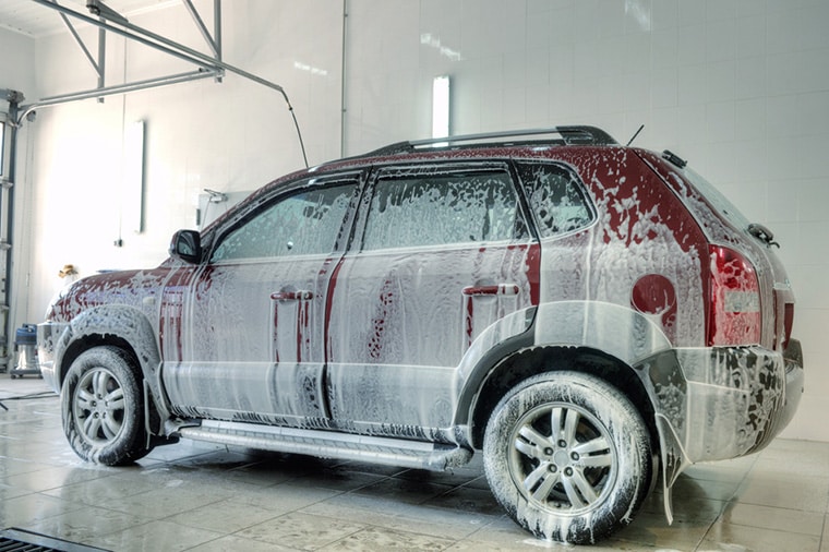 A red SUV is covered in soap suds during a car wash inside a garage. The vehicle is located on a tiled floor, and the garage is well-lit with bright white walls and ceiling lights.