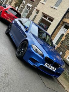 A blue BMW sports car, with license plate FM65 UCP, is parked on a street in front of a row of brick houses with steps and various colored doors. Another red car is parked in front of the BMW. The street is wet, suggesting recent rain.