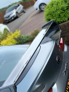 A close-up shot of the rear end of a gray sports car with a prominent carbon fiber spoiler. In the background, there are a few other parked cars and greenery along the edges of a paved driveway.