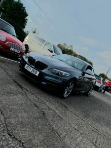 A dark gray BMW with the license plate "RF17 ZKD" is parked on a paved area next to other vehicles, including a red Mini Cooper and a white van. Trees and a fence are visible in the background under a clear sky.