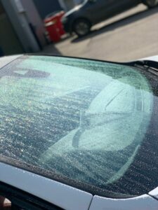 A car windshield covered in water droplets is captured in the sunlight, creating a glistening effect. The interior of the car, including the seats and dashboard, is partially visible through the glass. A red object and another parked car are seen in the background.