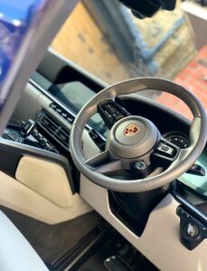 Close-up view of a luxury car's interior focused on the steering wheel with a Porsche logo. The dashboard features a digital display and sleek control buttons. The interior appears modern with leather and metallic accents, highlighted by natural light from the window.