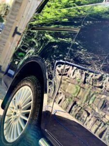 A close-up side view of a dark-colored, shiny car with reflections of nearby greenery and structures on its surface. The image highlights the car's polished exterior and rear wheel with decorative spokes and grooves on the tire.