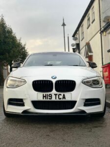 A front view of a white BMW car parked on a residential street, displaying license plate "H19 TCA". The background shows houses with satellite dishes and a red trash bin. The sky is overcast, suggesting a cloudy day.