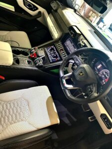 Interior of a luxury car featuring a high-tech steering wheel with a digital display, multiple control buttons, a gear shift, cup holders, and leather seats. The dashboard has a touchscreen interface and air vents, with the overall design showcasing modern aesthetics.