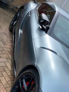 A sleek, metallic silver sports car is parked on a brick driveway. The vehicle features sharp lines and aerodynamic curves, with a glossy finish reflecting its surroundings. The close-up angle highlights the side mirror, wheel, and part of the door.