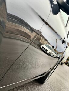 A close-up shot of a clean car's side panel with a clear reflection of a street, including houses and several parked cars. The shiny surface of the car emphasizes the reflections, creating a slightly distorted mirror-like effect.