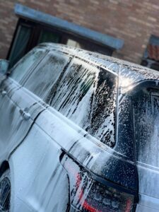 A black SUV covered in soap suds being washed. The vehicle is parked in a driveway near a brick building with windows. The soap suds are particularly visible on the rear and side windows of the vehicle.