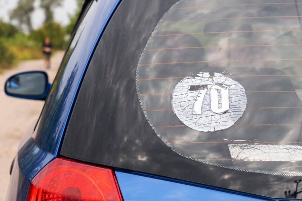 Close-up of a blue car's rear window sporting a peeling sticker with the number 70, hinting it might be time for some professional services. The background reveals a blurry outdoor scene filled with greenery and someone strolling in the distance.