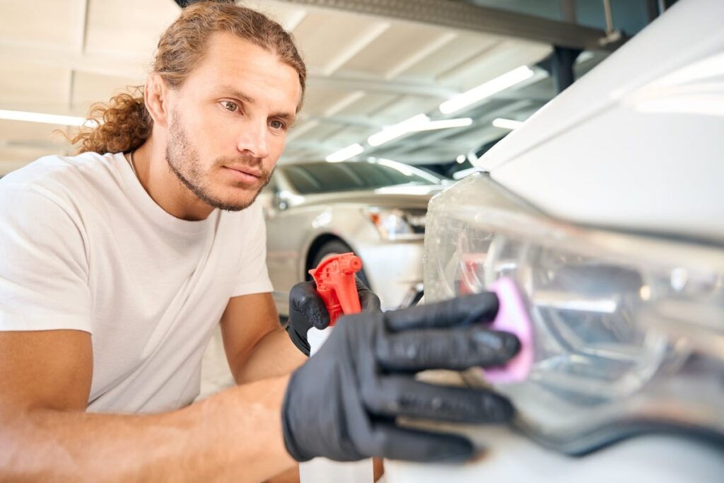 A person with long hair wearing gloves is polishing a car's headlight with a cloth. They're focused on detailing the vehicle, and there are cars parked in the background, inside a well-lit garage.