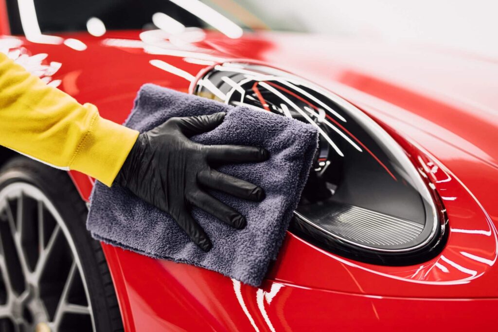 A person wearing a yellow sleeve and black gloves polishes the headlight of a shiny red sports car with a gray microfiber cloth. The car's sleek design and wheel are partially visible.